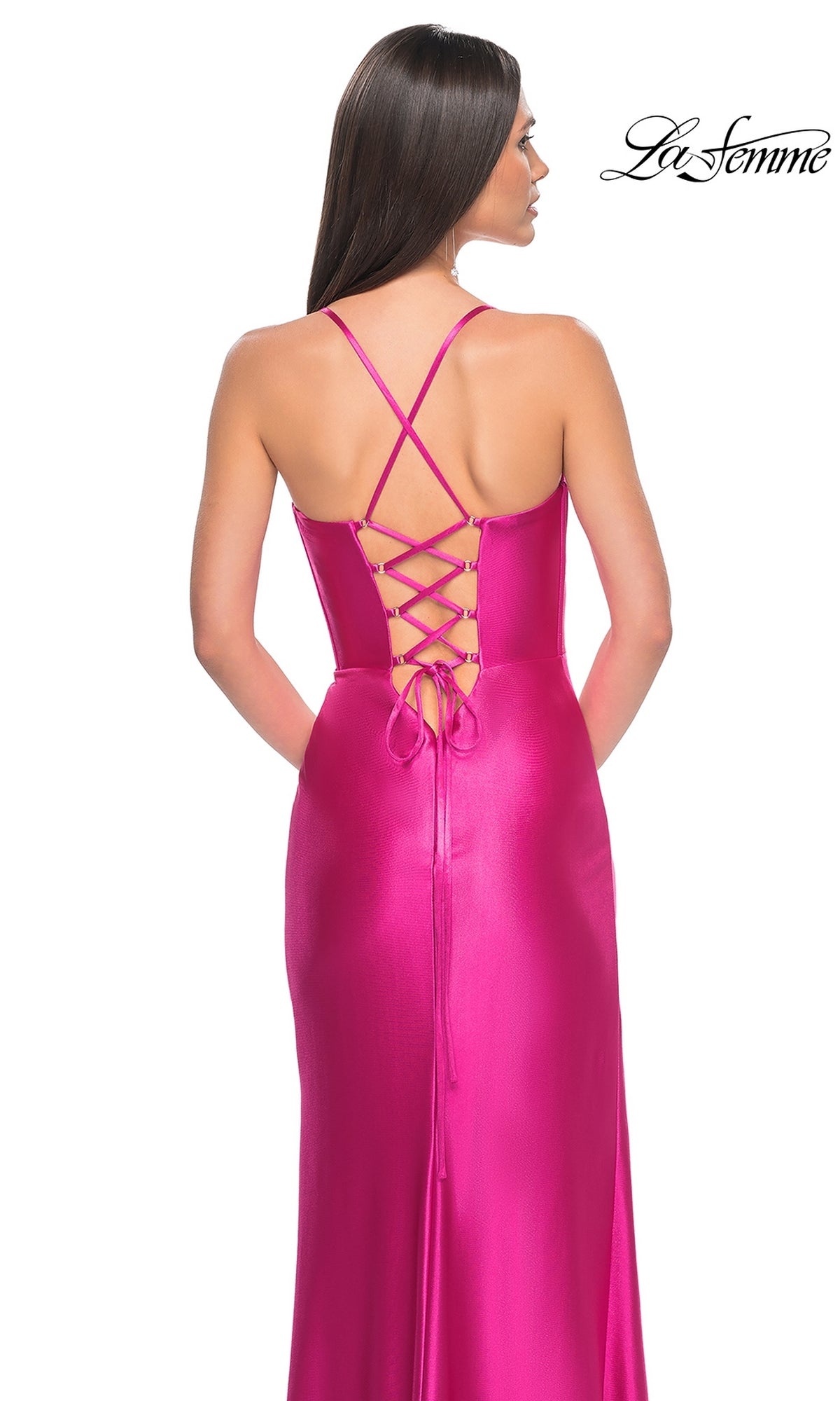 A woman stands with her back to the camera in the La Femme Bright Neon Long Prom Dress 32262, featuring shiny fuchsia fabric, crisscross straps, a corset-style bodice, and a lace-up back.